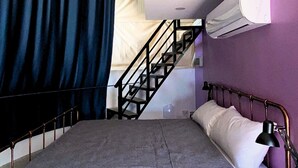A bedroom with a bed, a headboard, a nightstand, a lamp, a window with curtains, a staircase, and an air conditioning unit. - AmanDagat Beach Resort (San Felipe)