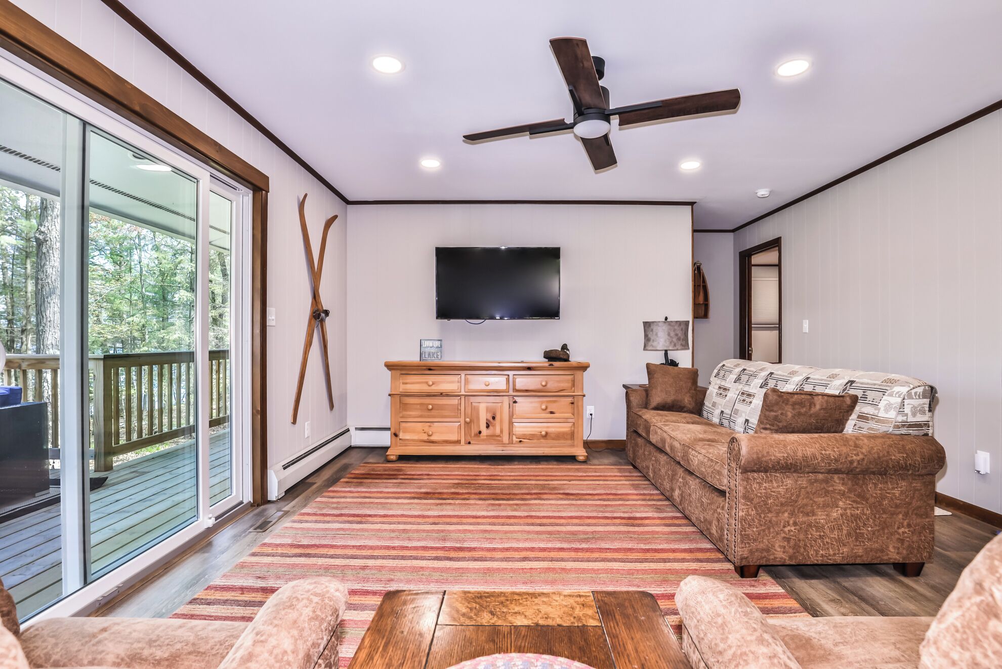 A spacious living room with a large window, a ceiling fan, a flat-screen TV, a wooden dresser, and a patterned sofa.