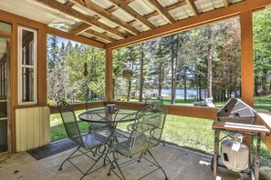 Une terrasse couverte avec une table et des chaises, un barbecue et une vue sur un lac entouré d’arbres.