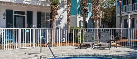 A pool area with a white fence, lounge chairs, and a building with blue shutters and palm trees.