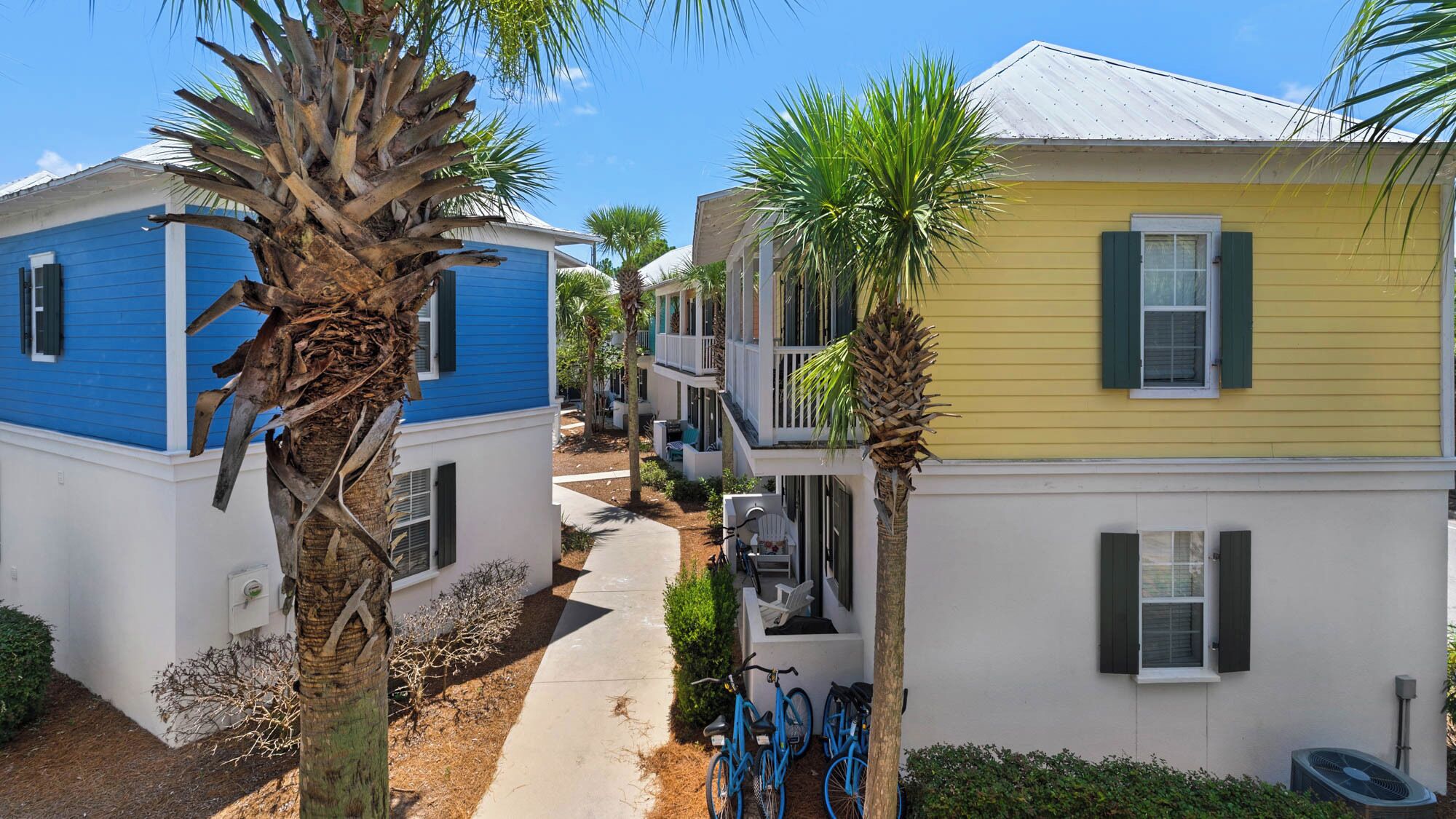 A narrow street with two-story houses, one blue and one yellow, with balconies and a palm tree in the foreground.