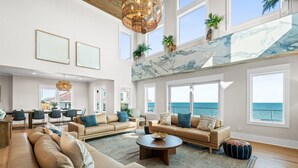 A spacious living room with a large window overlooking the ocean, a high ceiling with a unique light fixture, and a modern kitchen in the background. - Wavesong: Brand New Build, Beachfront, Private Pool, Pet Friendly, East End (St. George Island)