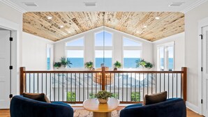 A room with a wooden ceiling, large windows, a balcony with a railing, and two blue chairs. - Wavesong: Brand New Build, Beachfront, Private Pool, Pet Friendly, East End (St. George Island)