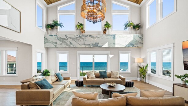 A spacious living room with a large window overlooking the ocean, a high ceiling with a chandelier, and multiple seating areas. - Wavesong: Brand New Build, Beachfront, Private Pool, Pet Friendly, East End (St. George Island)