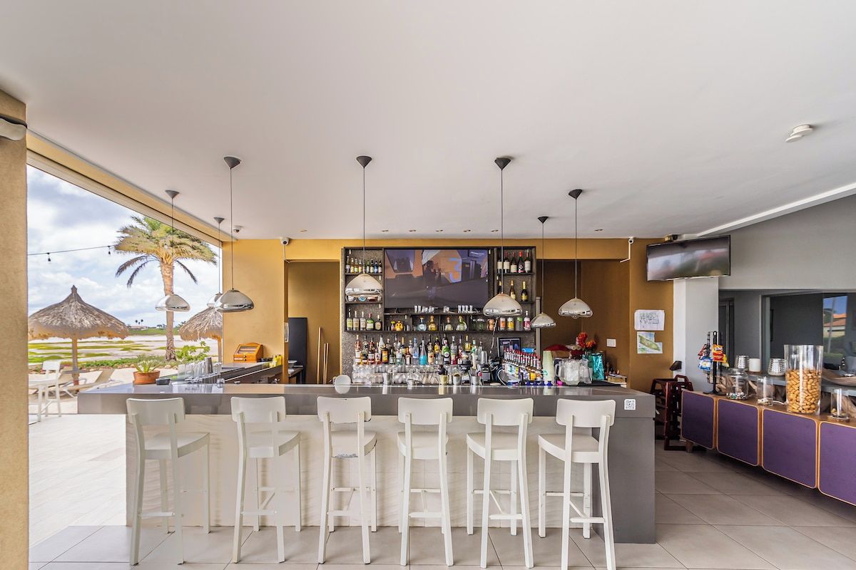 Una zona de bar con una barra alargada, varios taburetes y una variedad de botellas y vasos.