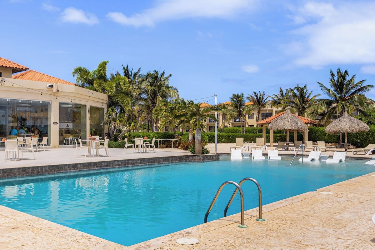Ein Swimmingpool mit Liegestühlen und Sonnenschirmen, umgeben von Palmen und einem Gebäude mit einem Ziegeldach.