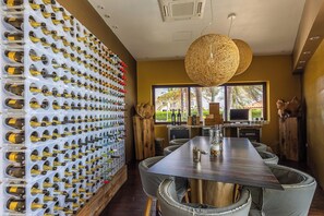 A dining area with a long table, cushioned chairs, a wine rack with bottles, and a large woven light fixture. - Stunning 3BR Oasis w Private Pool Patio near Beach (Noord)