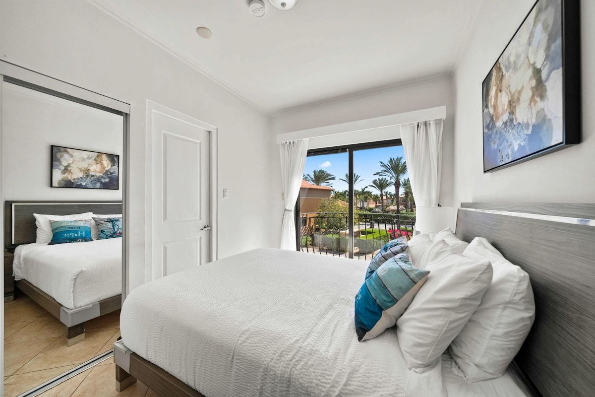Une chambre d’hôtel avec un grand lit, un lit plus petit et un balcon offrant une vue sur les palmiers.