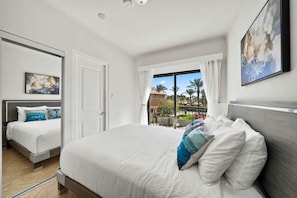 Quarto de hotel com uma cama grande, uma cama menor e uma varanda com vista para palmeiras.