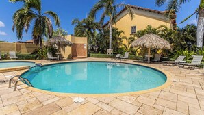 Une piscine entourée de palmiers et d’un bâtiment, avec des chaises longues et des parasols.
