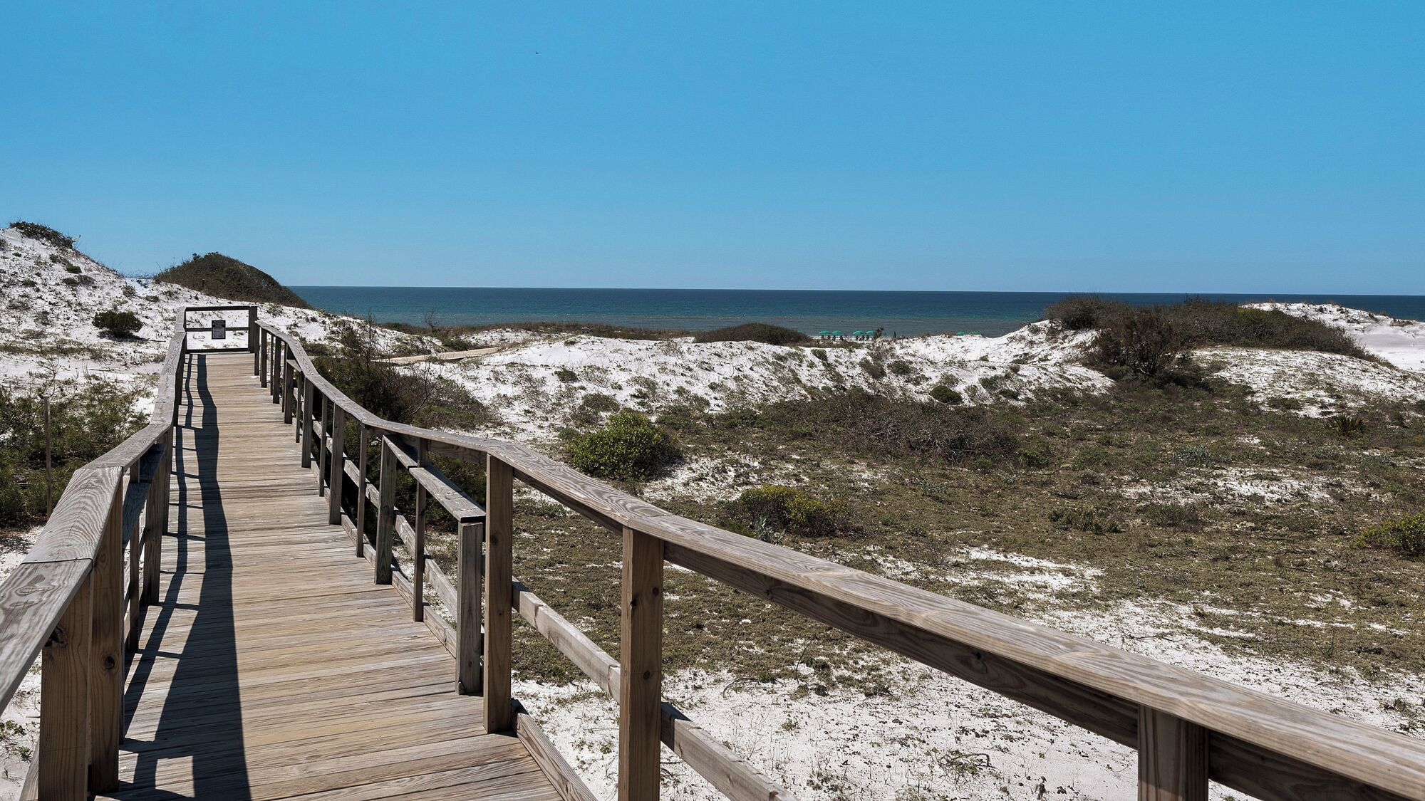 Une promenade en bois qui mène à une plage bordée de dunes, sous un ciel bleu et dégagé.