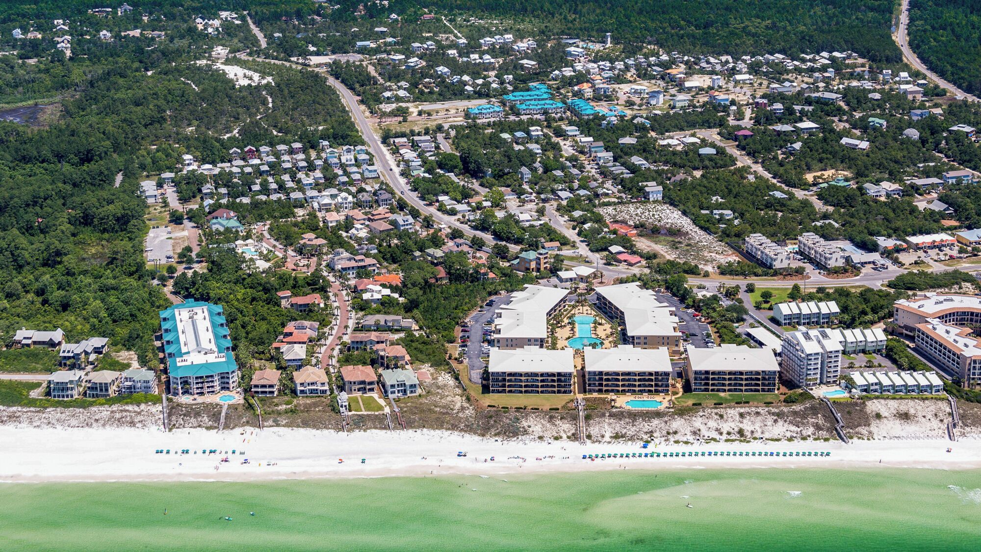 An aerial view of a coastal residential area with houses, a beach, and a large building with a swimming pool.