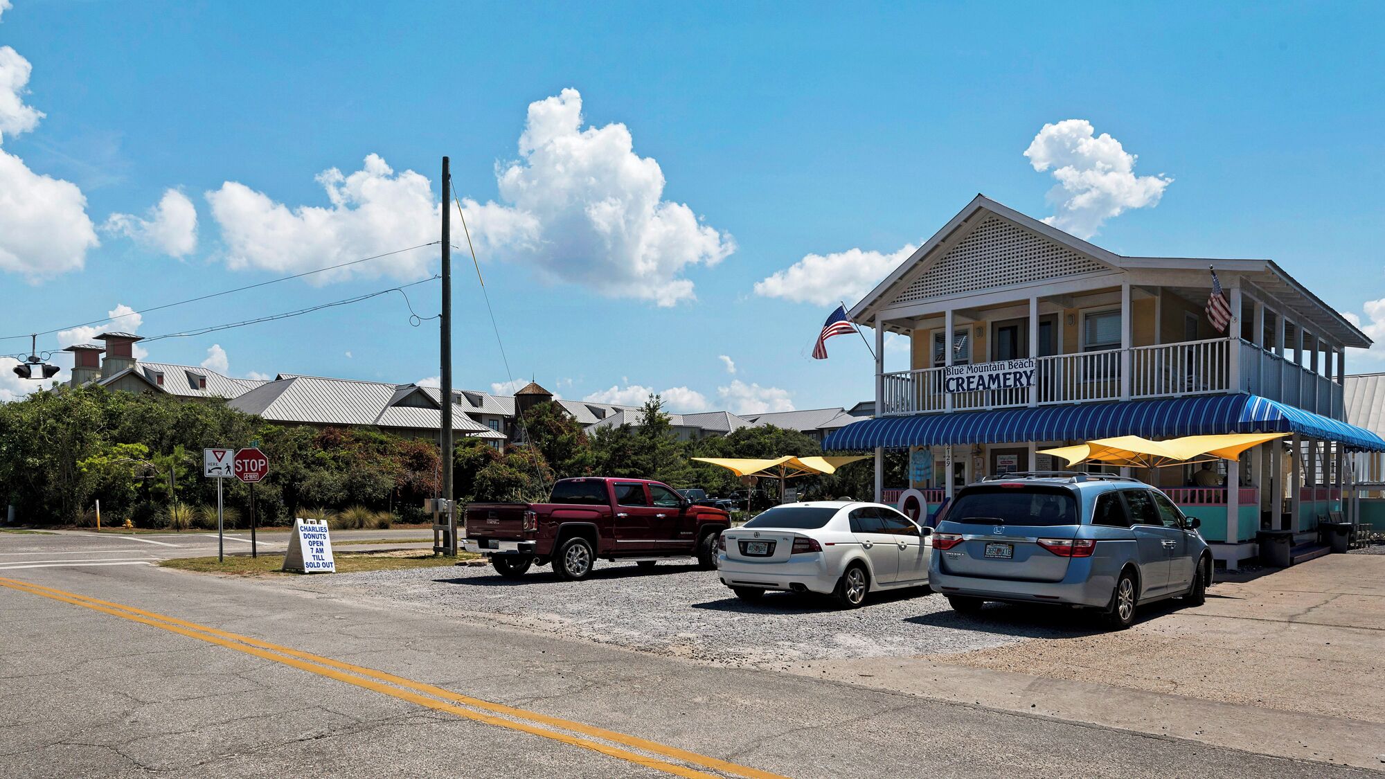A creamery with a porch and umbrellas, a stop sign, and a truck parked on the side of the road.