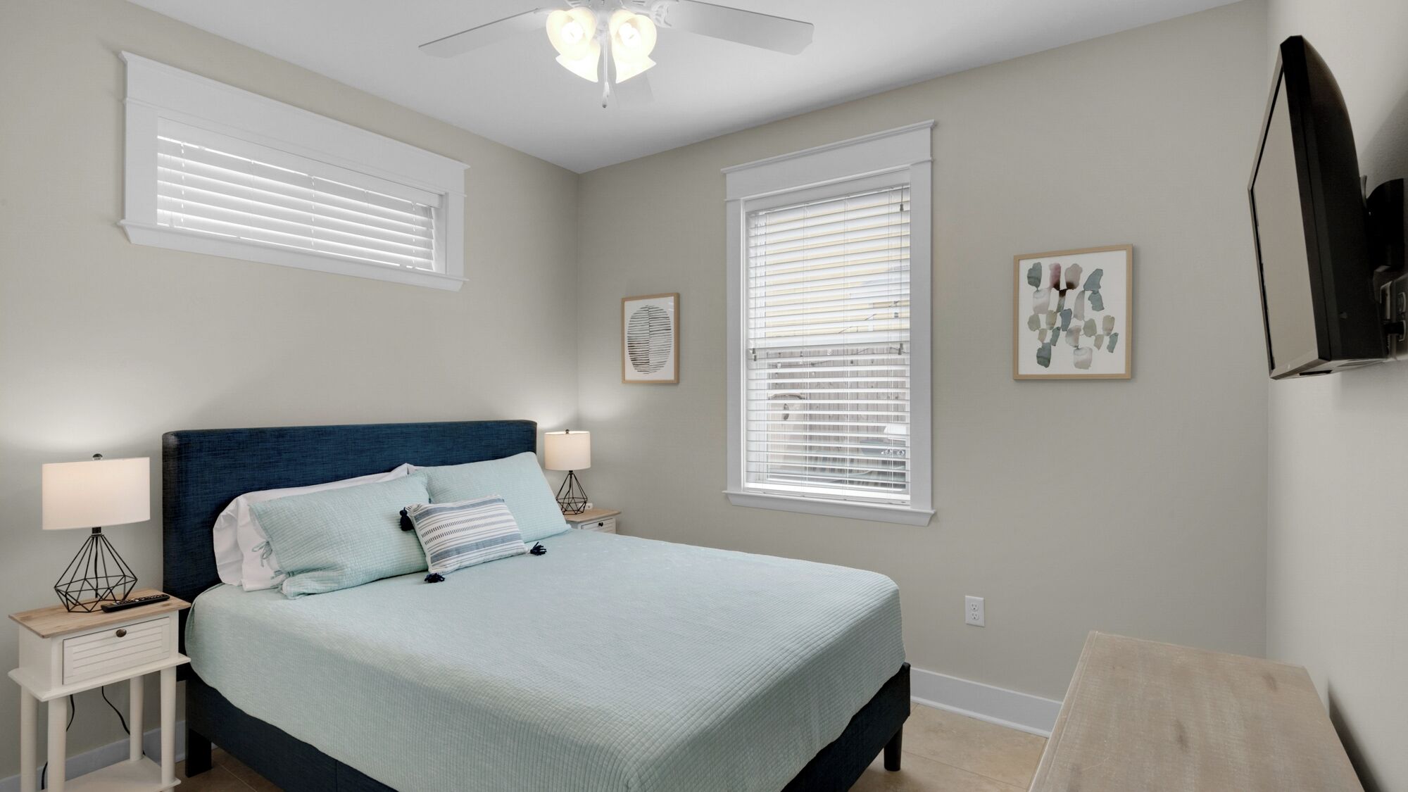 A bedroom with a bed, two bedside tables with lamps, a ceiling fan, and two windows with blinds.