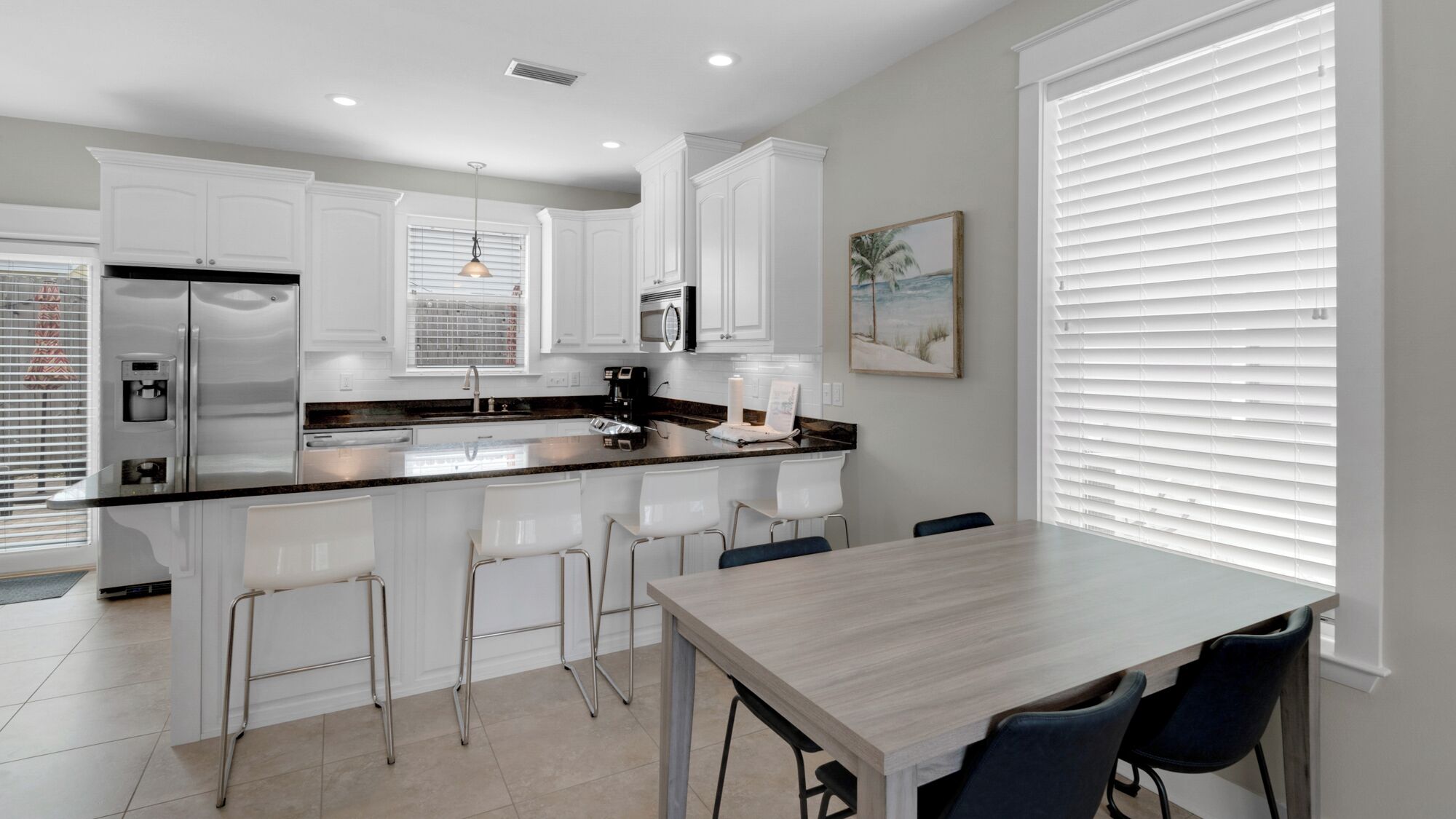 Una cocina moderna con armarios blancos, una isla central con taburetes, una mesa de comedor y un ventanal con persianas.