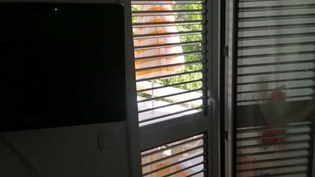 Habitació amb una finestra gran amb persianes horitzontals, televisió penjada a la paret i zona exterior visible amb vegetació.