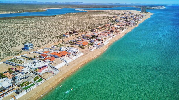 Vista aérea de un complejo turístico costero con una extensa playa, aguas azules cristalinas y una combinación de edificios frente al mar y en el interior.