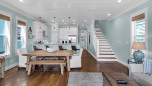 Una cocina amplia con armarios blancos, una mesa de comedor de madera e iluminación colgante. Una escalera conduce a un segundo piso.