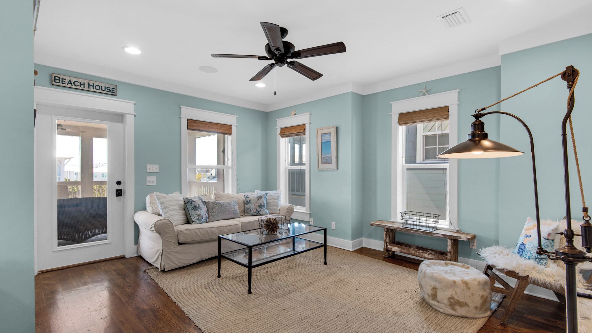 A living room with a sofa, coffee table, and a ceiling fan. There is a sign that reads "Beach House" on the door.