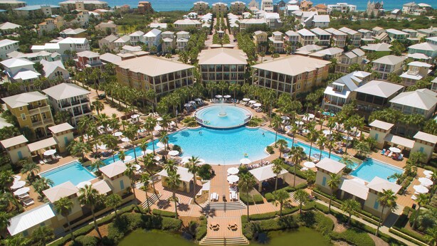 Vista aérea de un resort con varias edificaciones, una piscina central y palmeras.