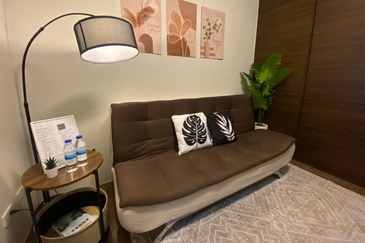 A room with a brown sofa, a small table with a lamp, a potted plant, and wall art.