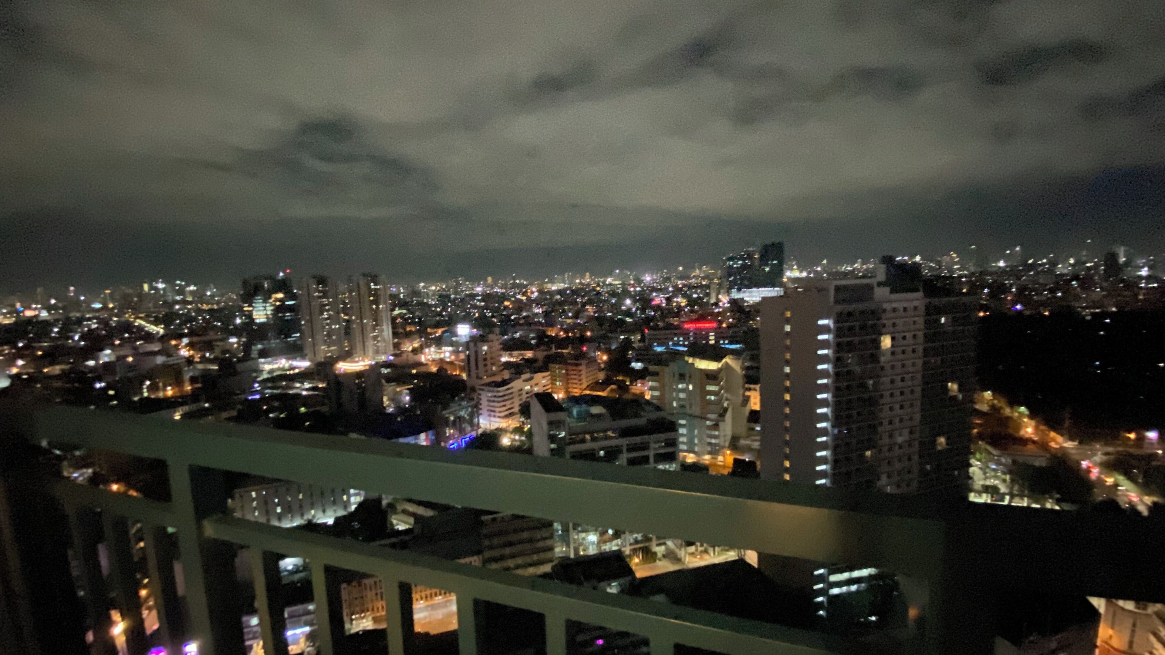 Une vue nocturne d’une ville, prise depuis un point d’observation élevé, avec des bâtiments illuminés et un ciel nuageux.