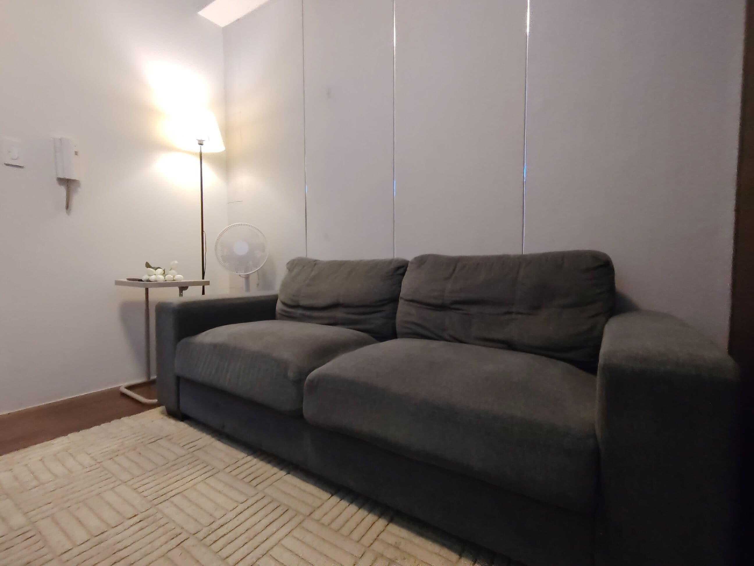 Een kamer met een grijze bank, een kleine tafel met een lamp, en een staande lamp.