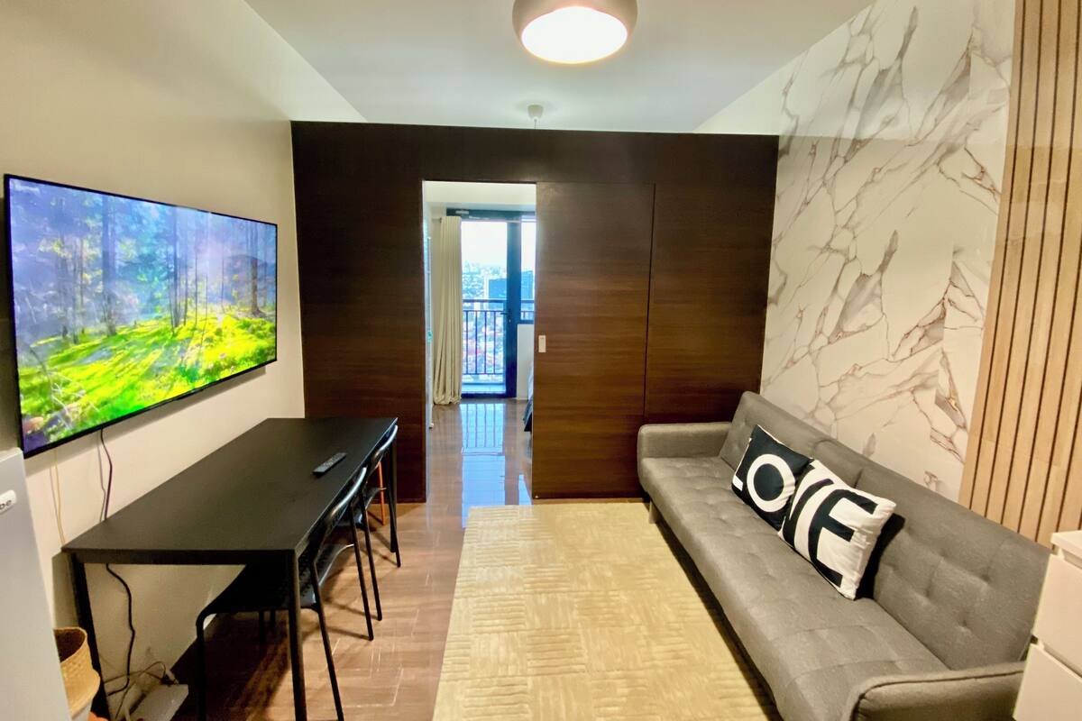 A modern living room with a flat-screen TV, a sofa with a 'LOVE' pillow, a dining table with chairs, and a view into the apartment.