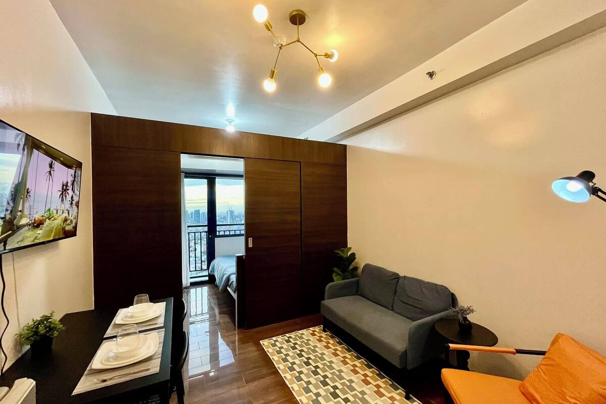 A modern living room with a sofa, a dining table set, a patterned rug, and a view of the city through a large window.