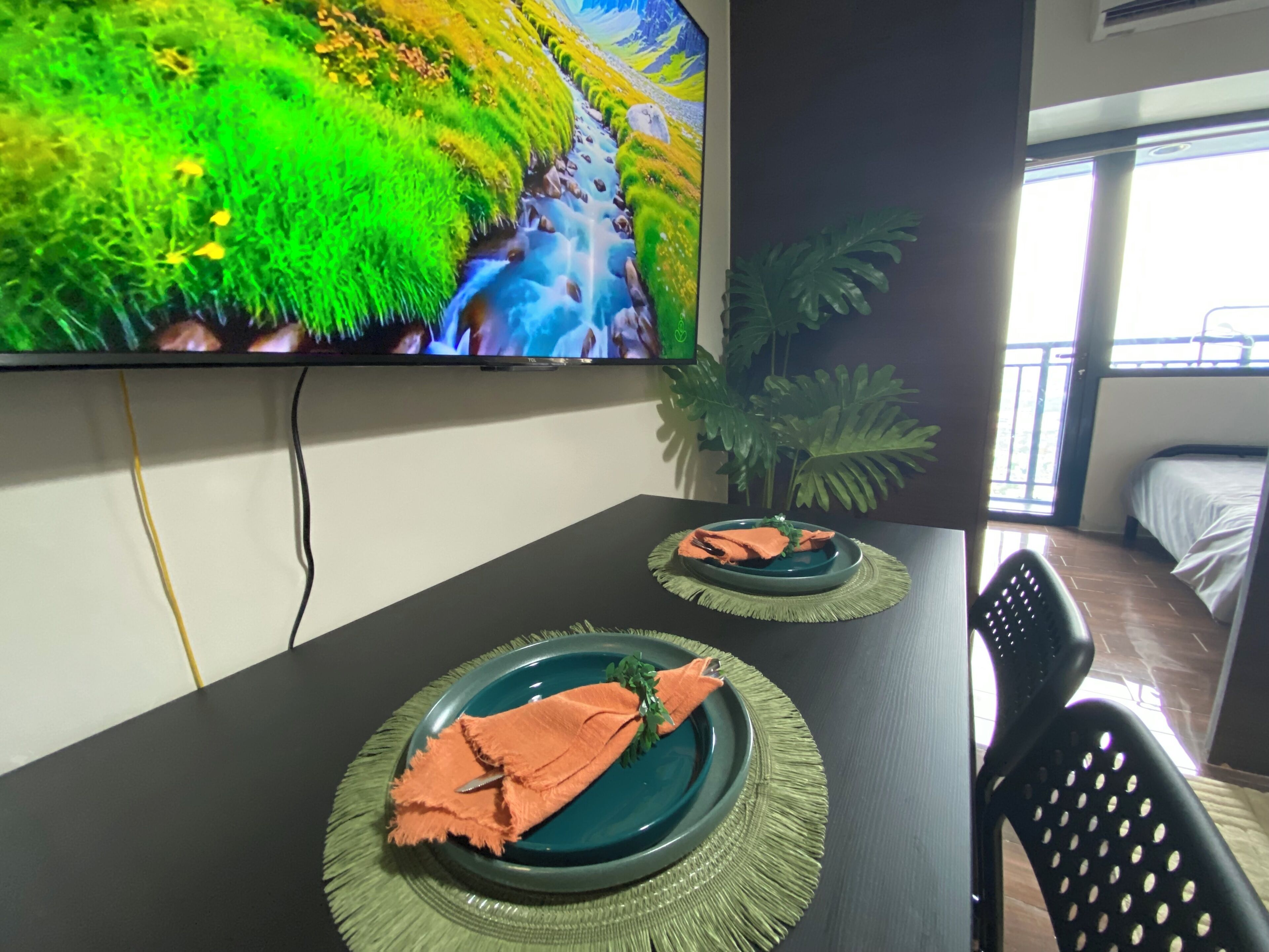 A dining table with two plates, each with a green placemat and an orange napkin. A large TV mounted on the wall displays a nature scene.