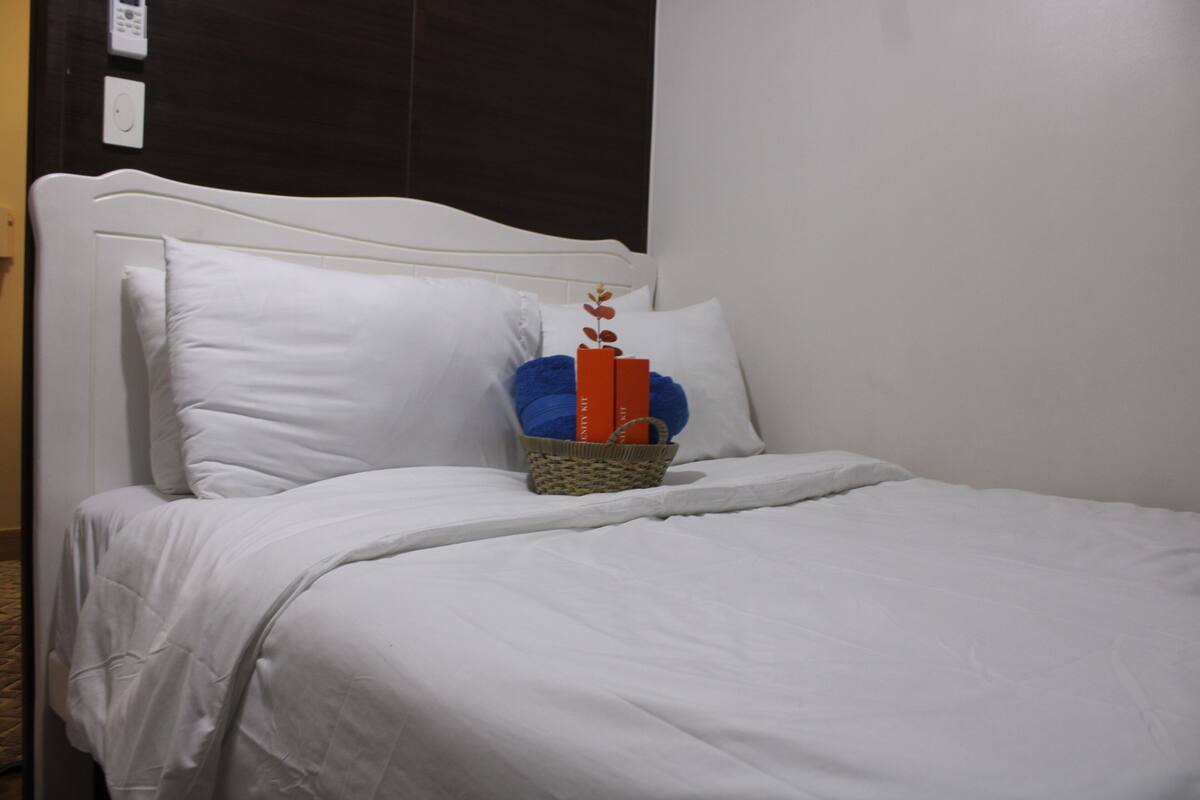A neatly made bed with white linens, a basket with books, and a wall-mounted thermostat.