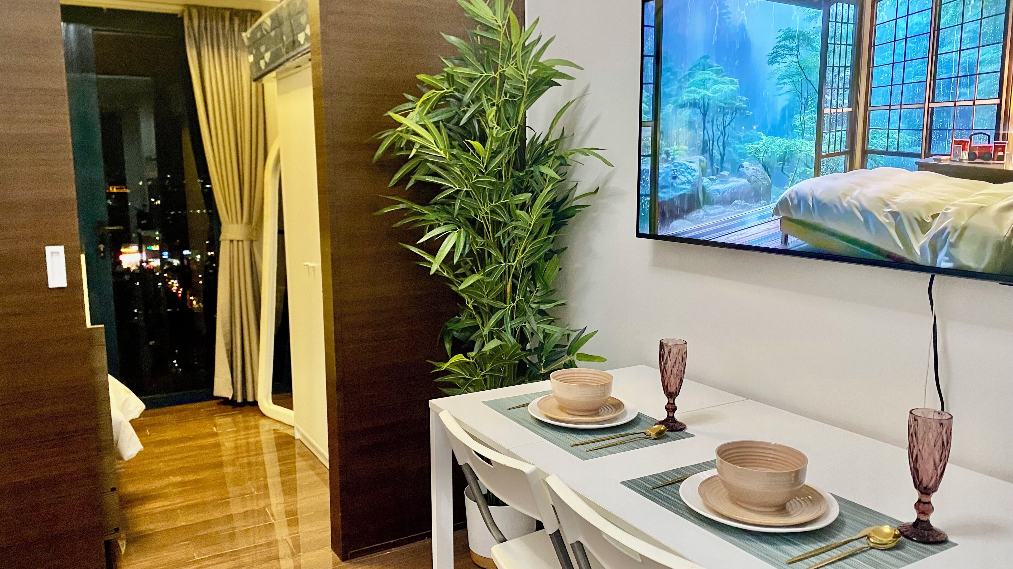 A hotel room with a dining table set for two, a large potted plant, and a TV mounted on the wall displaying a scenic view.