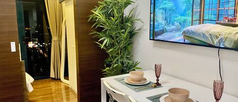 A hotel room with a dining table set for two, a large potted plant, and a TV mounted on the wall displaying a scenic view.