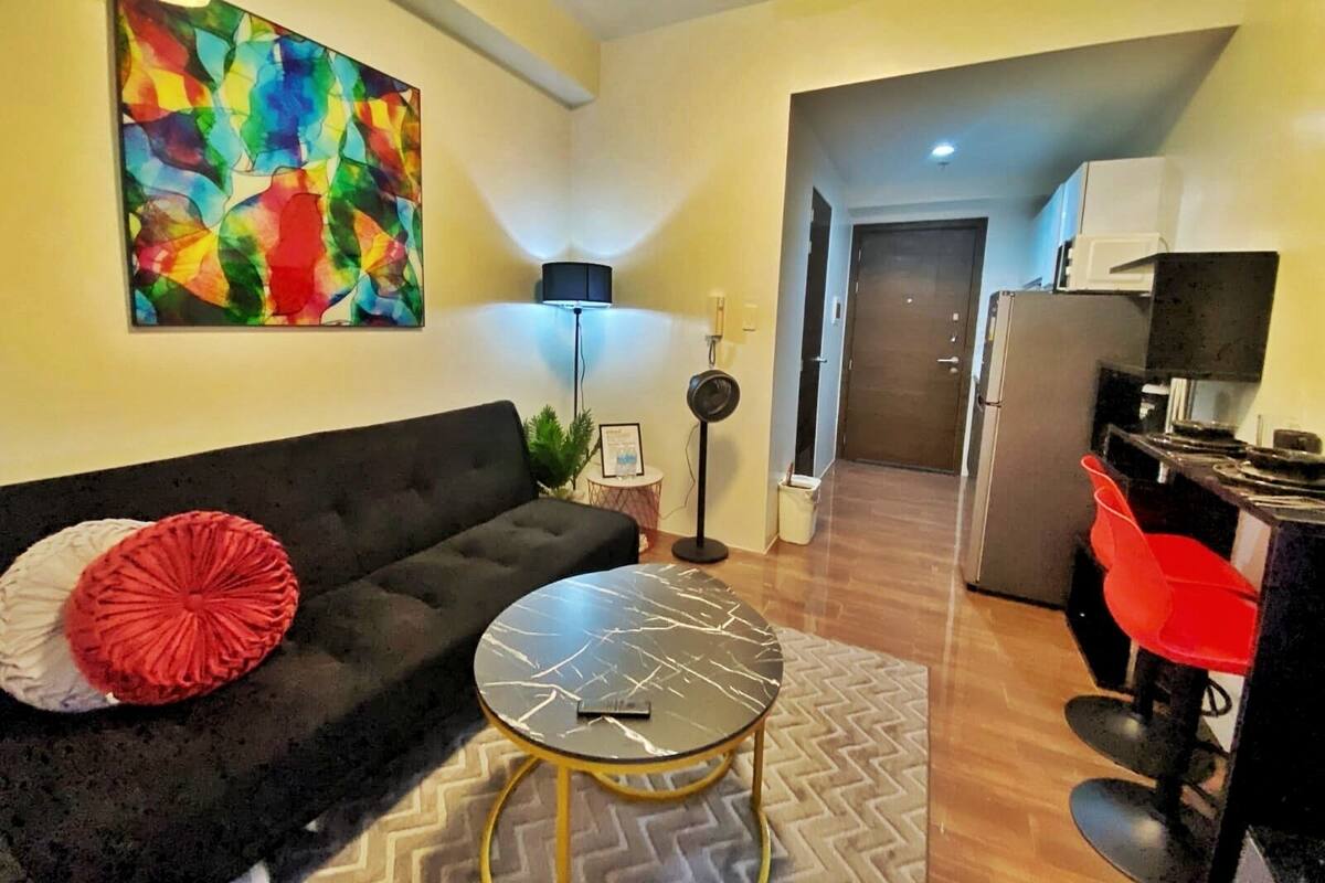 A living room with a black sofa, a round coffee table, a colorful abstract painting, a lamp, and a kitchen area with a refrigerator and red chairs.