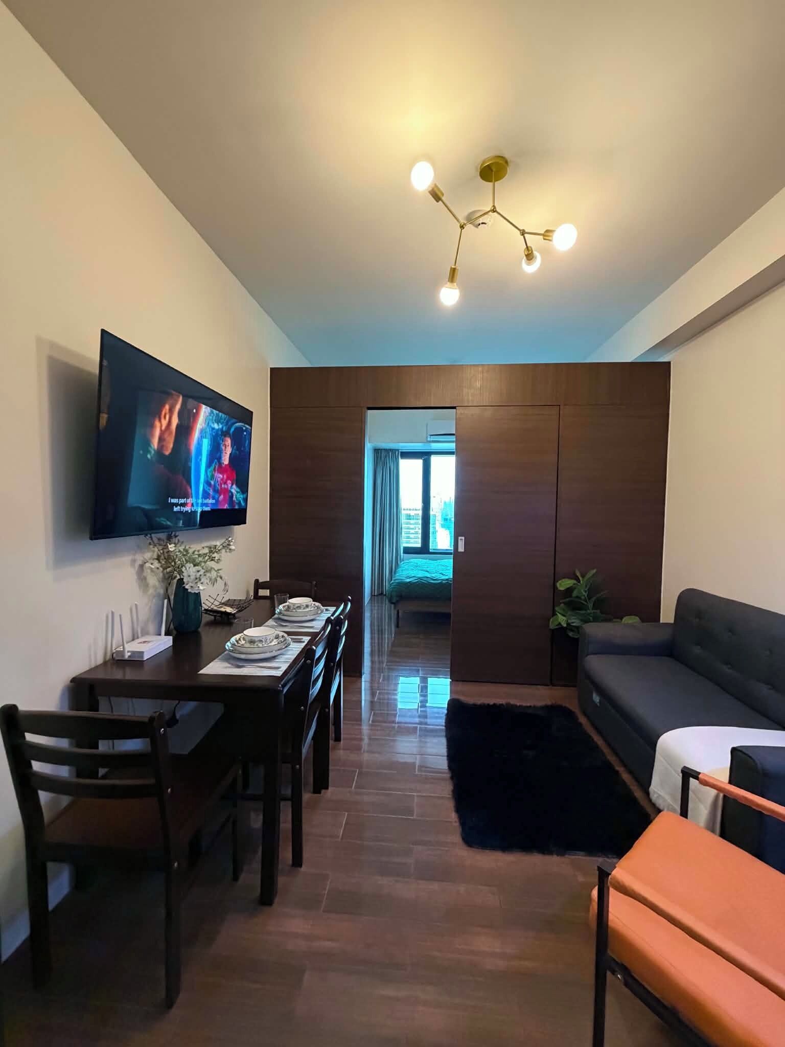Uma sala de estar moderna com uma TV de tela plana, uma área de refeições com mesa e cadeiras, um sofá e um lustre.