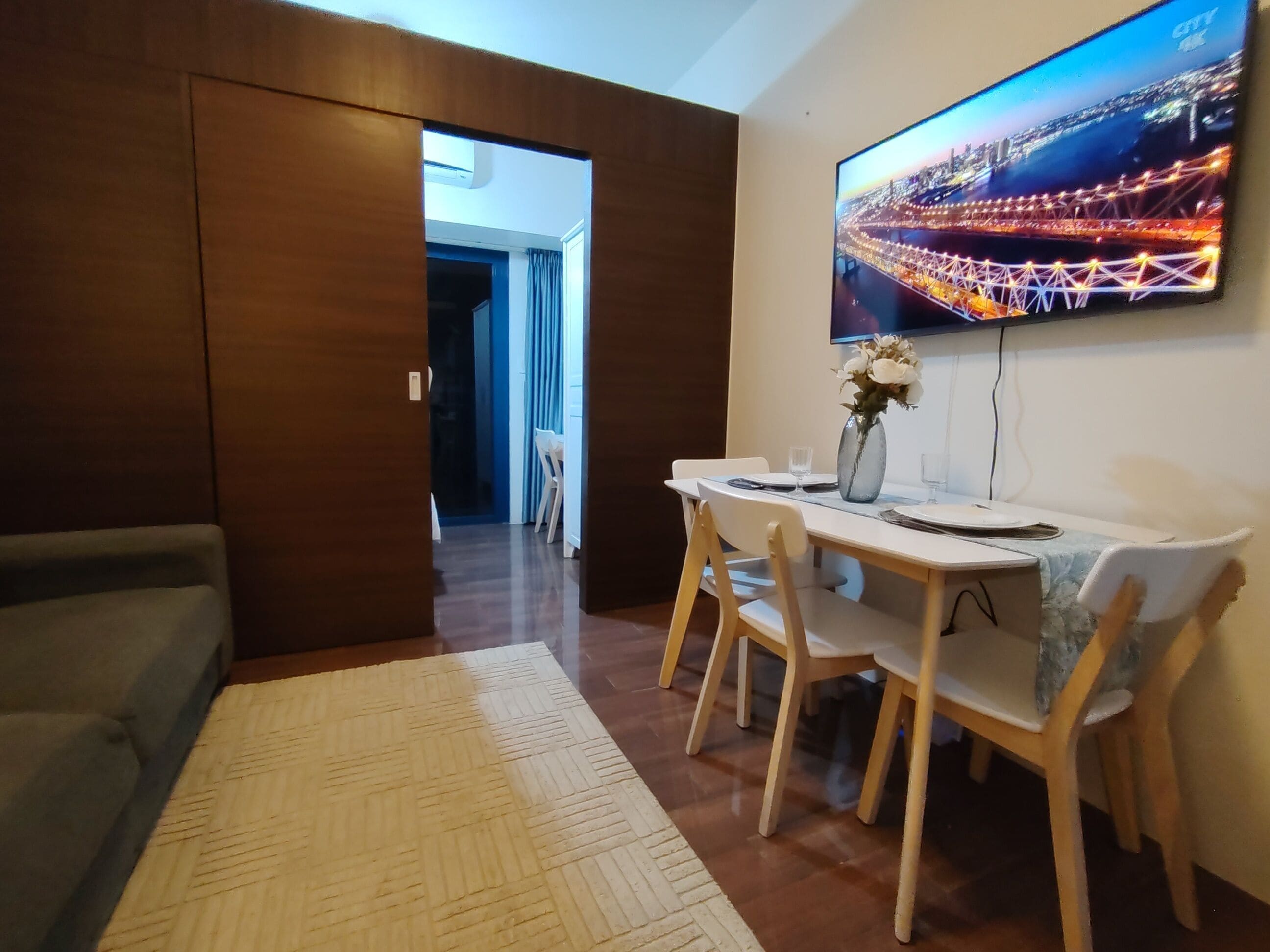 A dining area with a table set for four, a vase with flowers, and a TV displaying a cityscape.