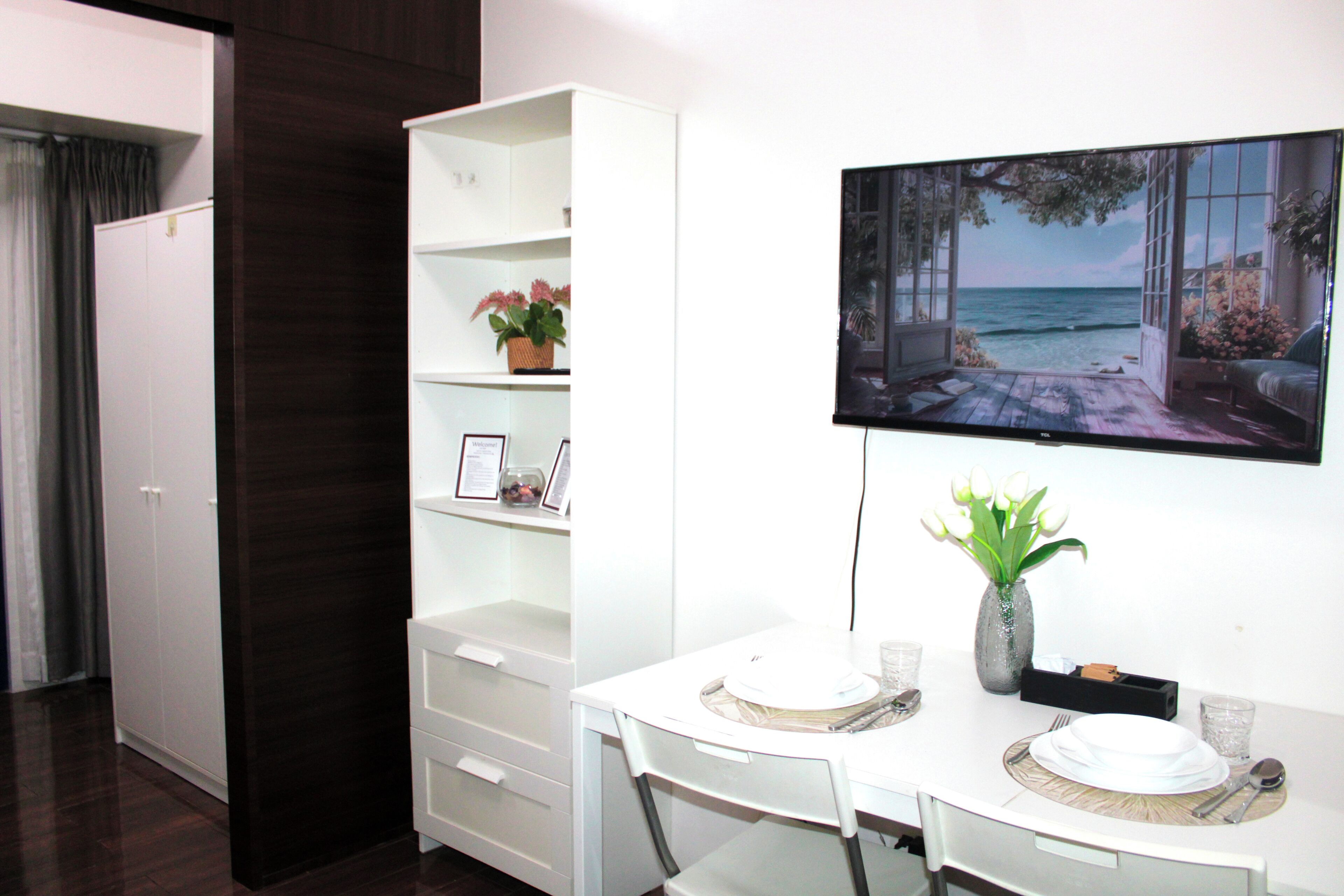 A modern room with a white dining table, chairs, a flat-screen TV mounted on the wall, and a white shelving unit with decorative items.