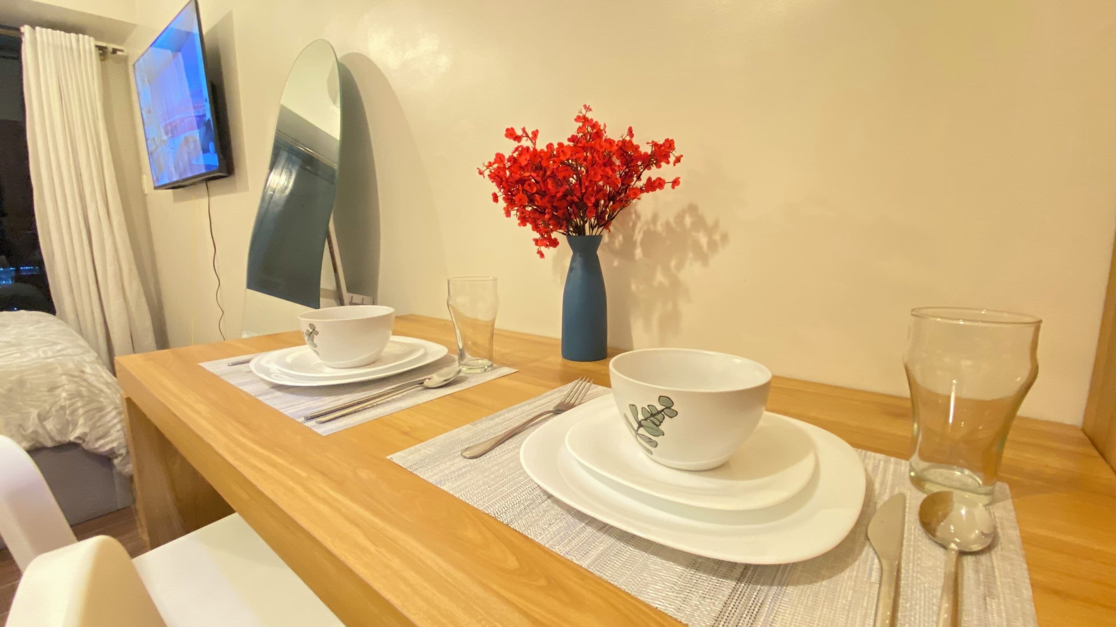 Une table à manger bien dressée, avec de l’argenterie blanche, des assiettes et des bols, un vase rempli de fleurs rouges et un téléviseur à écran plat fixé au mur.