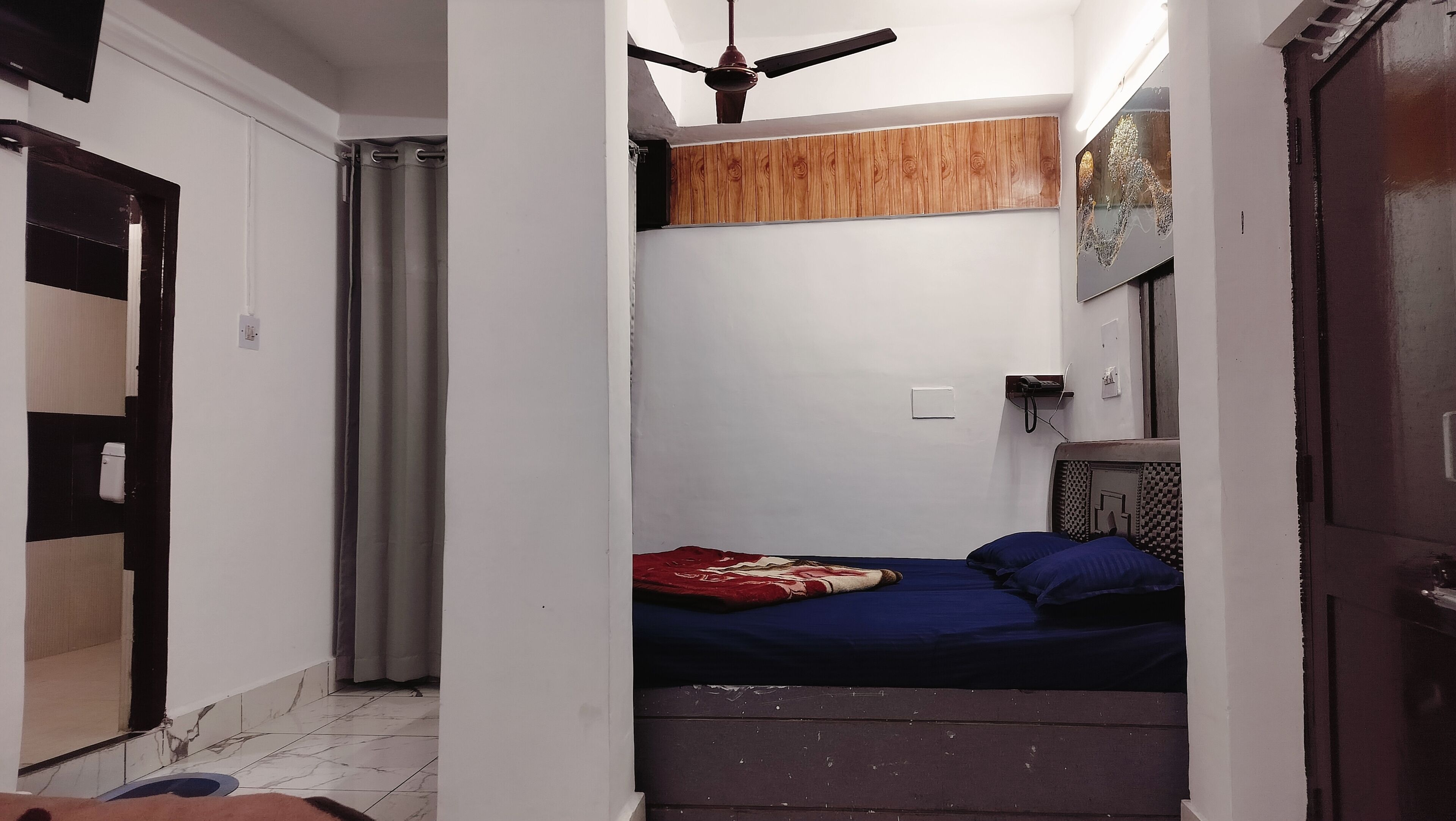 A small, narrow room with a bed, a ceiling fan, a wooden panel above the bed, and a door on the right.