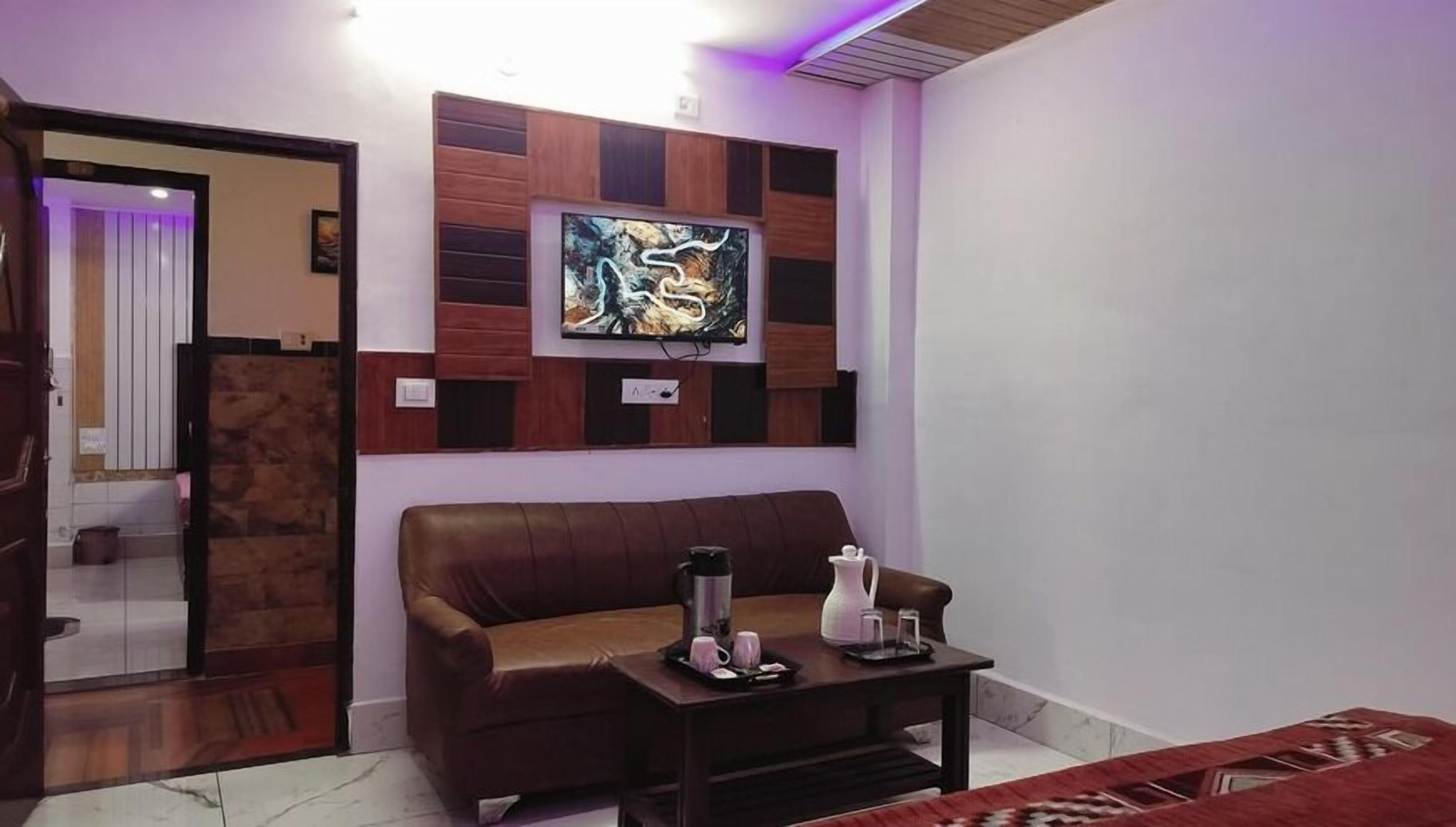 A living room with a brown leather sofa, a coffee table with a kettle and cups, and a wall-mounted TV.