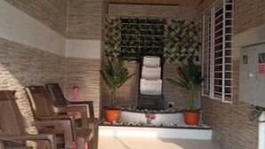 Un espace extérieur abrité, aménagé avec des chaises et des plantes en pot, un mur en pierre et une fenêtre ornée d’une grille métallique.