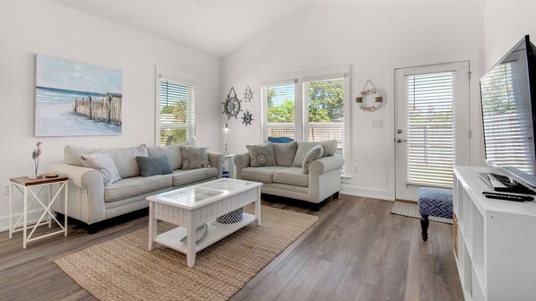 A spacious living room with two sofas, a coffee table, and a TV. There are large windows with blinds, a rug, and a painting on the wall. - Beautiful Coastal Chic Home for 9 + Neighborhood Pool in Panama City Beach (Panama City Beach)