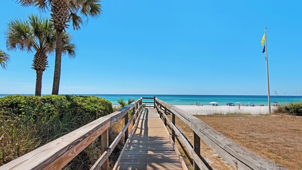 A wooden boardwalk leading to a beach with palm trees and a flag. - Beautiful Coastal Chic Home for 9 + Neighborhood Pool in Panama City Beach (Panama City Beach)