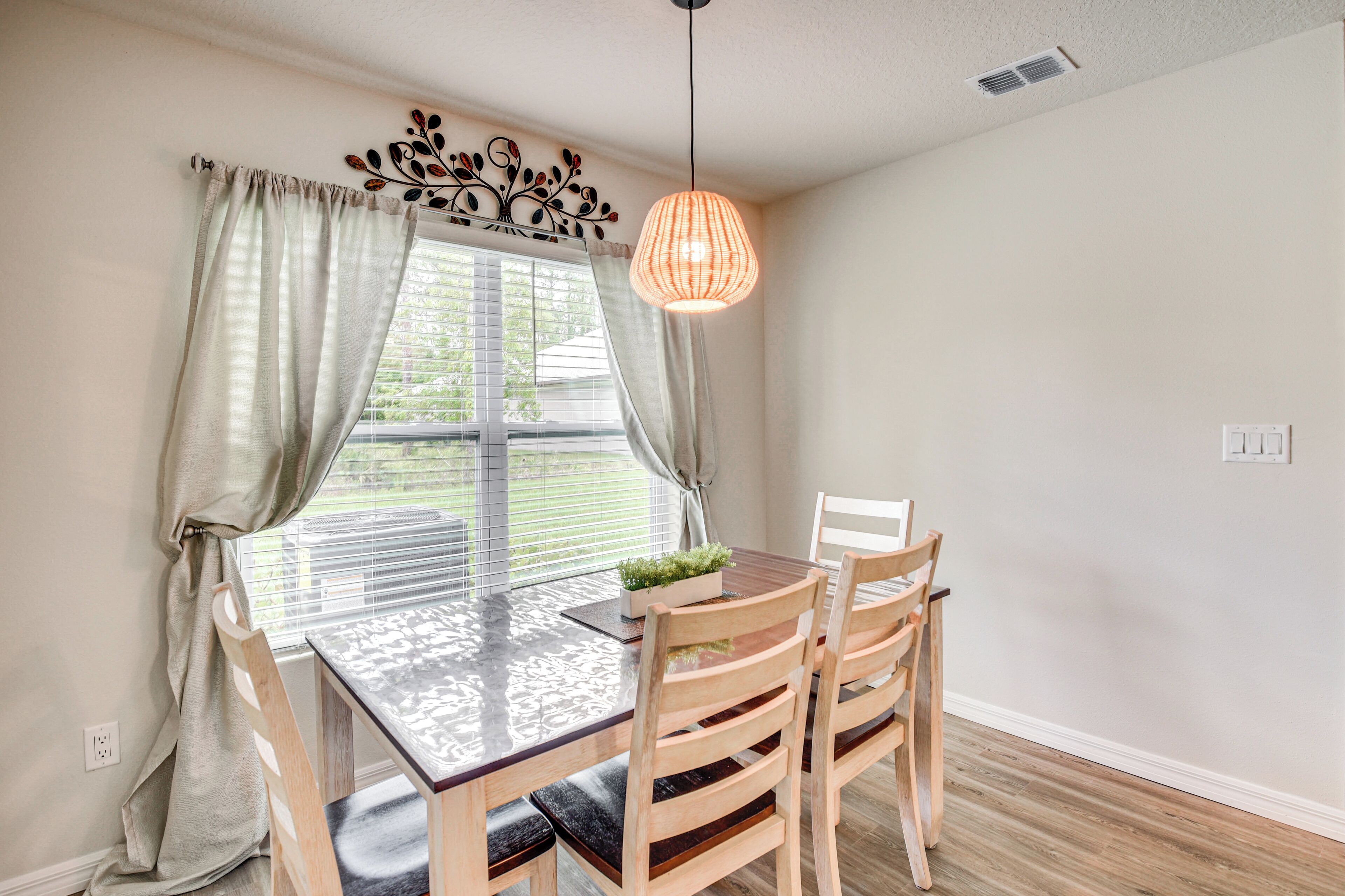 A dining area with a wooden table, four chairs, a hanging light fixture, and a window with curtains.