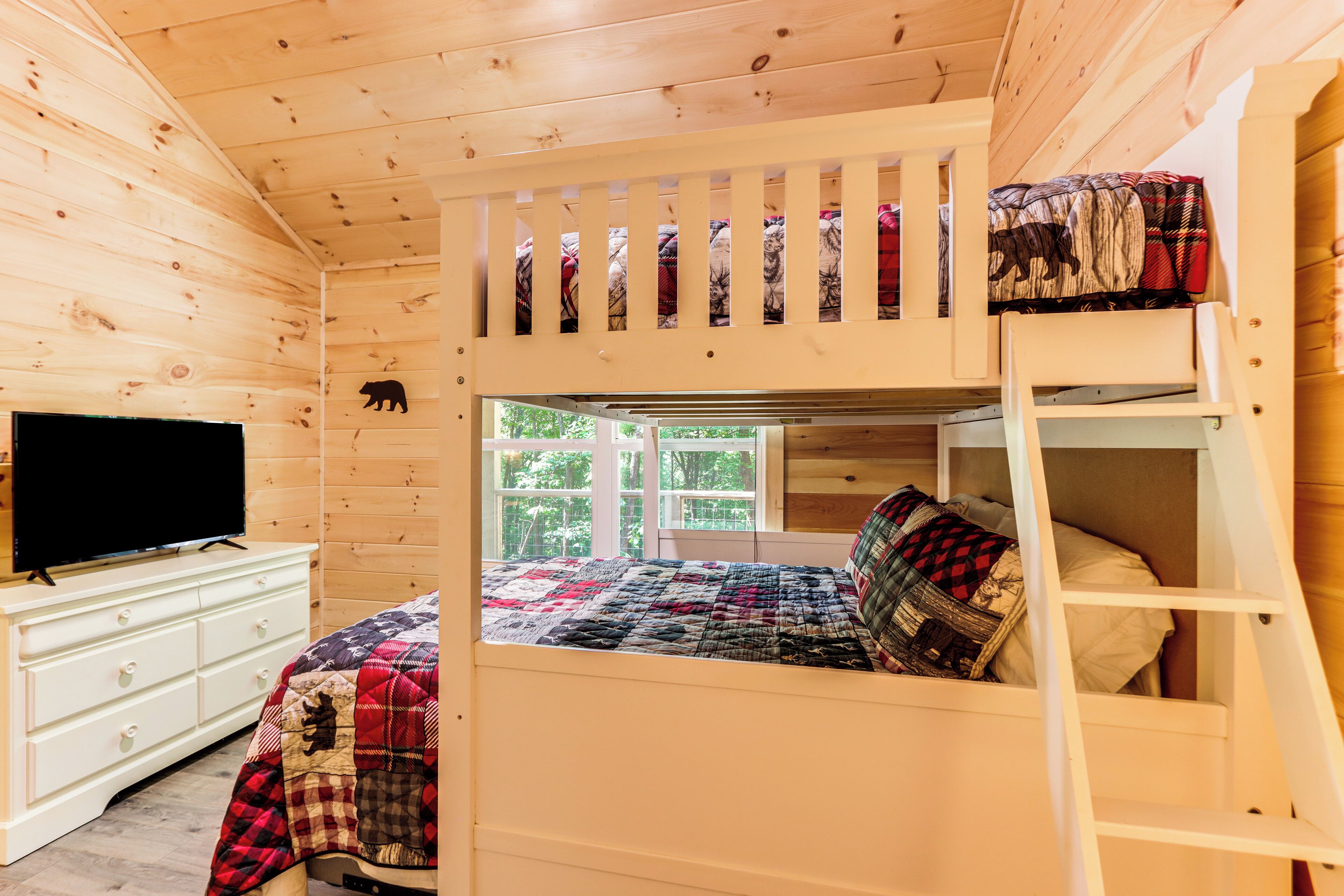 Une chambre avec des lits superposés, une télévision posée sur une commode, une fenêtre donnant sur des arbres et une découpe d’ours accrochée au mur.