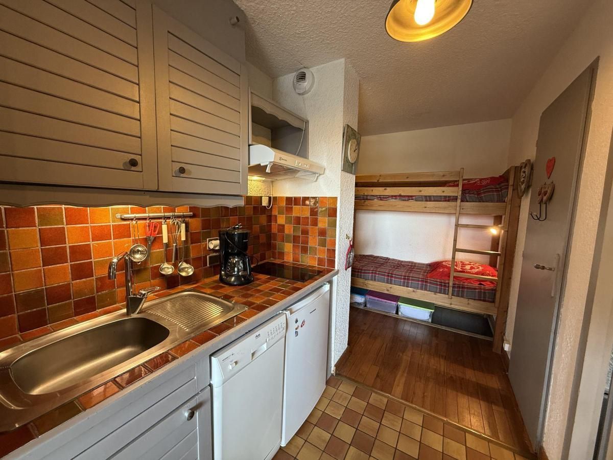 A compact kitchen with a sink, stove, and white cabinets. A bunk bed with a red blanket is visible in the background.