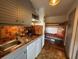 A compact kitchen with a sink, stove, and white cabinets. A bunk bed with a red blanket is visible in the background.