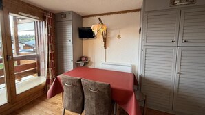 A room with a wooden floor, a table with a red tablecloth, two chairs, a television on the wall, a radiator, and a large window with curtains. - 150m from ski slopes, 2nd floor, view ski slopes, balcony, parking, tv, ski locker, 29m² (Les Saisies)