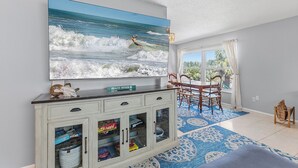 Uma sala de estar com uma televisão grande fixada na parede, uma área de jantar com mesa e cadeiras, e um armário com gavetas.