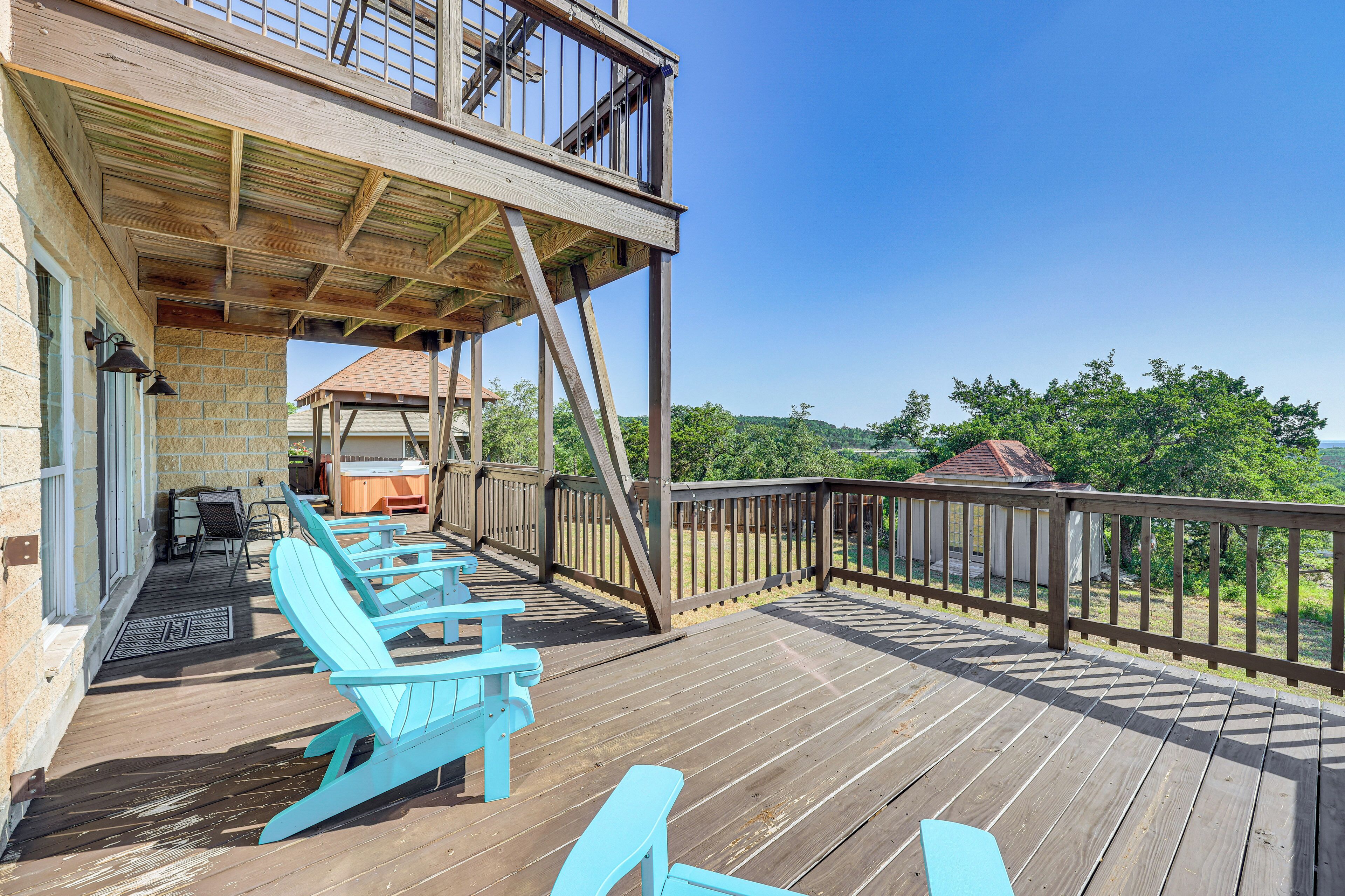 A wooden deck with blue Adirondack chairs, a covered area with a railing, and a view of a house and trees in the background.